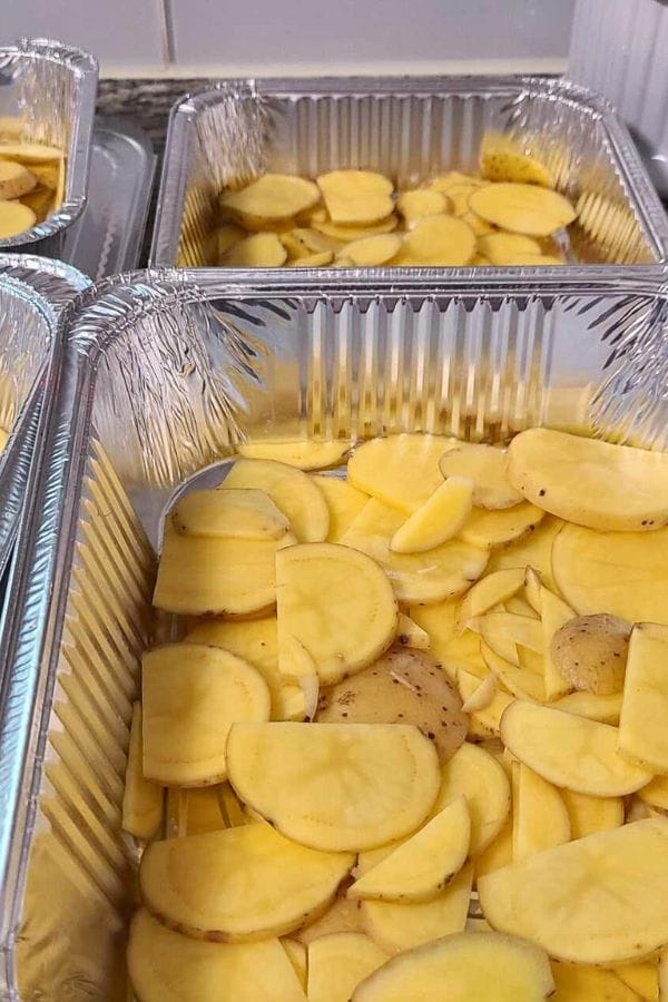Sliced potatoes, reminiscent of a cheeseburger and fries casserole, are meticulously arranged in multiple aluminum trays.
