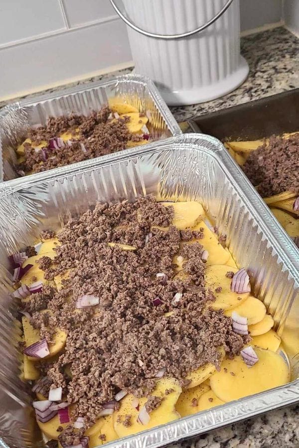 Aluminum trays filled with sliced potatoes, ground meat, chopped red onions, and seasoning create a delightful cheeseburger and fries casserole scene on the kitchen counter.