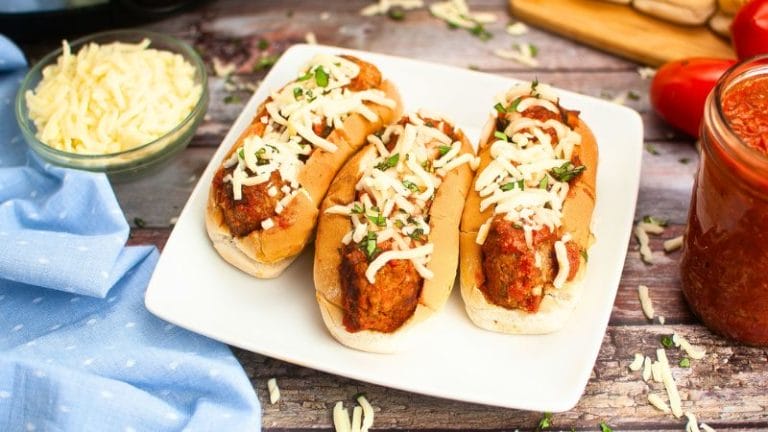 A trio of meatball subs topped with shredded cheese and chopped herbs on a white plate. Nearby, a jar of sauce, a bowl of cheese, and a blue cloth complete the cozy spread.