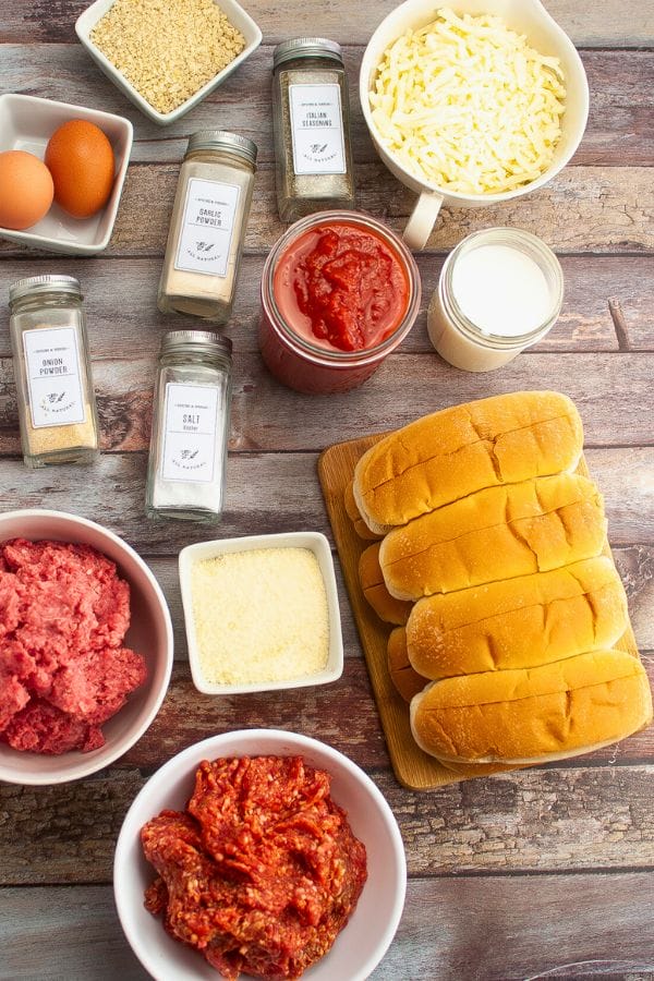 Ingredients for making meatball subs, including ground meat, marinara sauce, spices, eggs, cream, shredded cheese, grated cheese, and breadcrumbs on a wooden surface. Hoagie rolls complete the ensemble for a hearty meal