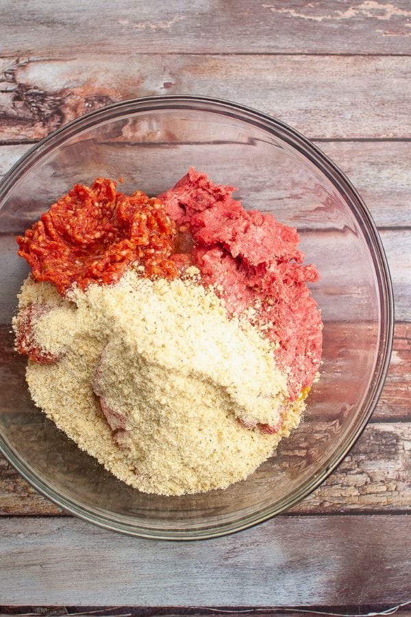 A glass bowl with ground meat, red sauce, and breadcrumbs sits on a wooden surface,