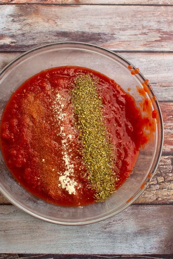 A glass bowl with tomato sauce, topped with oregano, garlic powder, and paprika, sits on a wooden surface