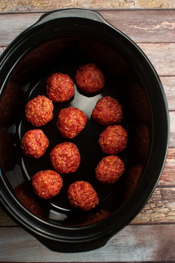 A black slow cooker containing twelve evenly spaced meatballs rests on a wooden surface, 
