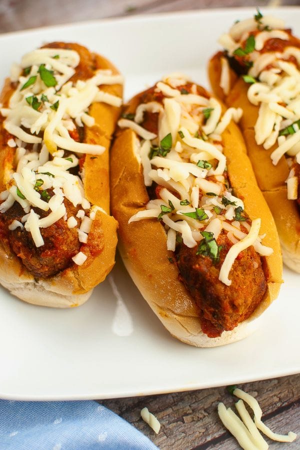 Three meatball subs topped with melted cheese and fresh herbs on a white plate, offering comfort