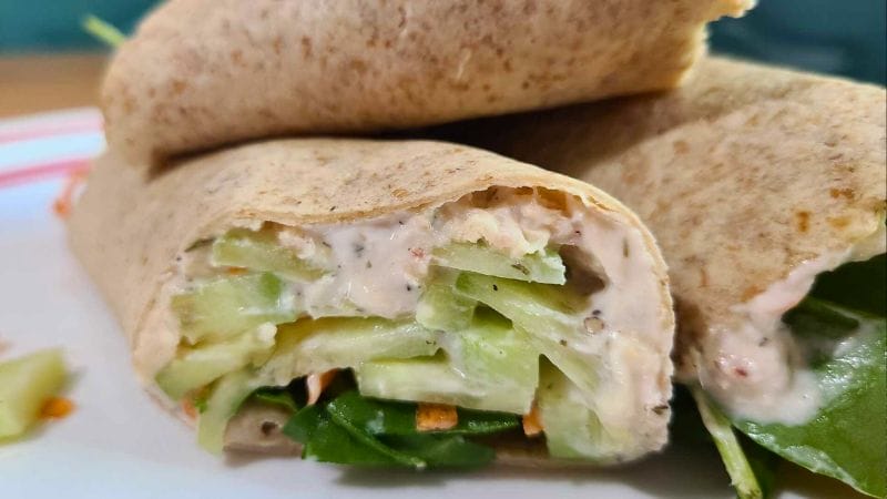 Close-up of a healthy tuna wrap stuffed with sliced cucumbers, greens, and creamy sauce, displayed on a white plate.