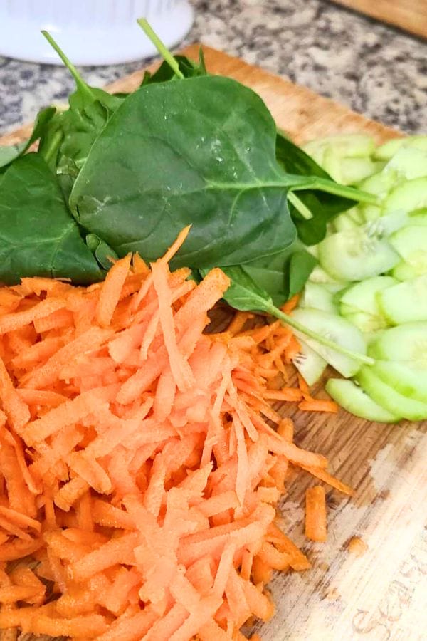 Chopped spinach, sliced cucumbers, and shredded carrots on a wooden cutting board, ready to be wrapped into a healthy tuna wrap.