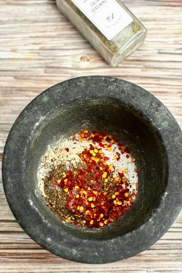 A stone mortar contains mixed spices and red pepper flakes, reminiscent of an Olive Garden Dressing Dry Mix, with a spice jar in the background on a wooden surface.