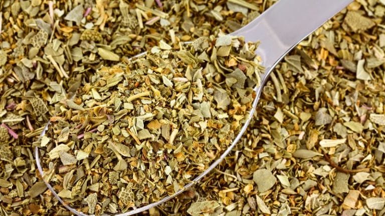 A close-up of a metal spoon filled with dried mixed herbs, surrounded by more herbs