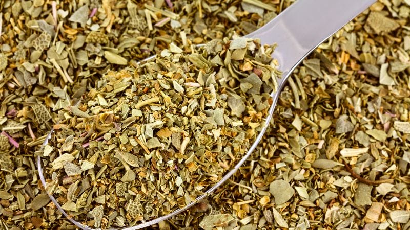 A close-up of a metal spoon filled with dried mixed herbs, surrounded by more herbs