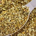 A close-up of a metal spoon filled with dried mixed herbs, surrounded by more herbs