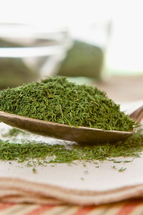 A spoonful of dried dill on a cloth napkin with a bowl in the blurred background.