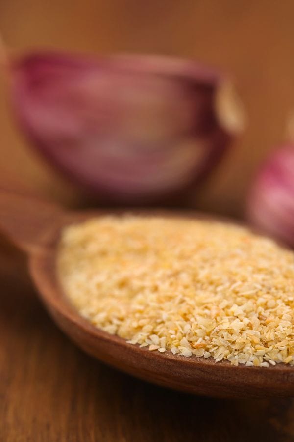 A wooden spoon filled with granulated garlic takes center stage, while purple garlic cloves form a blurred backdrop