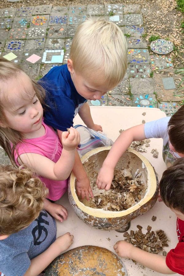Children are gathered around a table, examining a large bowl filled with dried plant material, as they eagerly discuss how to make drums. The area is paved with colorful, mosaic-like tiles, adding a vibrant backdrop to their creative exploration.