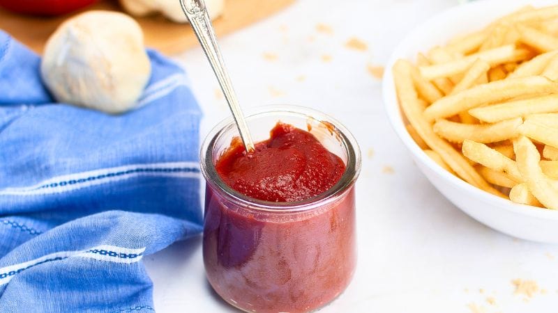 A jar of ketchup with a spoon next to a blue cloth and a bowl of fries
