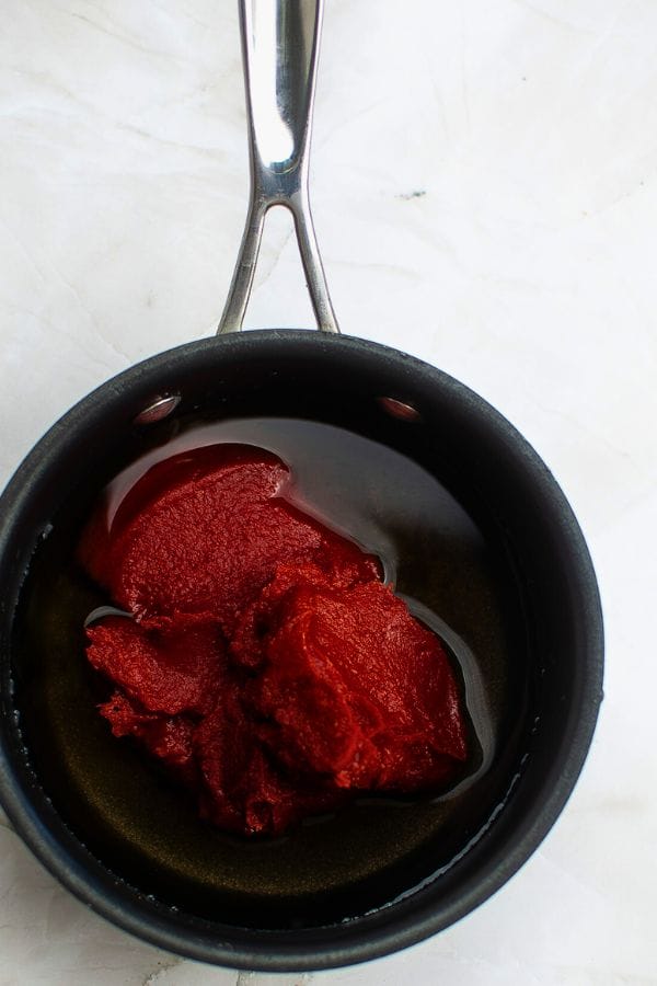 A black saucepan containing tomato paste and oil sits on a white surface