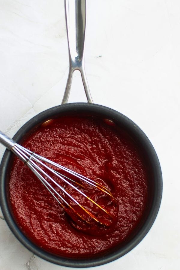 A saucepan of rich red tomato sauce with a metal whisk resting inside, positioned on a white surface