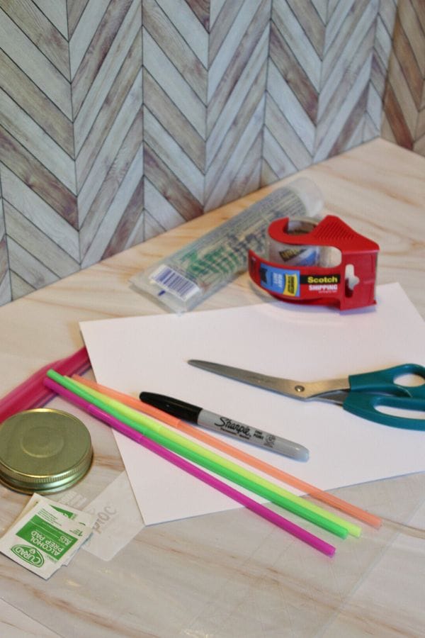 A creative array of crafting supplies awaits on the table: scissors, tape, straws, a Sharpie, paper, and gum packets. Nearby sits a mason jar lid next to sprinkle squish bags ready to spark your imaginative projects.