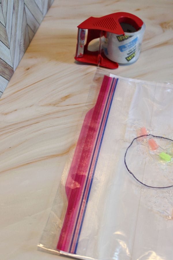 A Sprinkle Squish Bag with a vibrant zipper rests on the table, accompanied by a red tape dispenser and a roll of tape. The clear plastic design perfectly complements its colorful counterparts, adding flair to your organizing essentials.