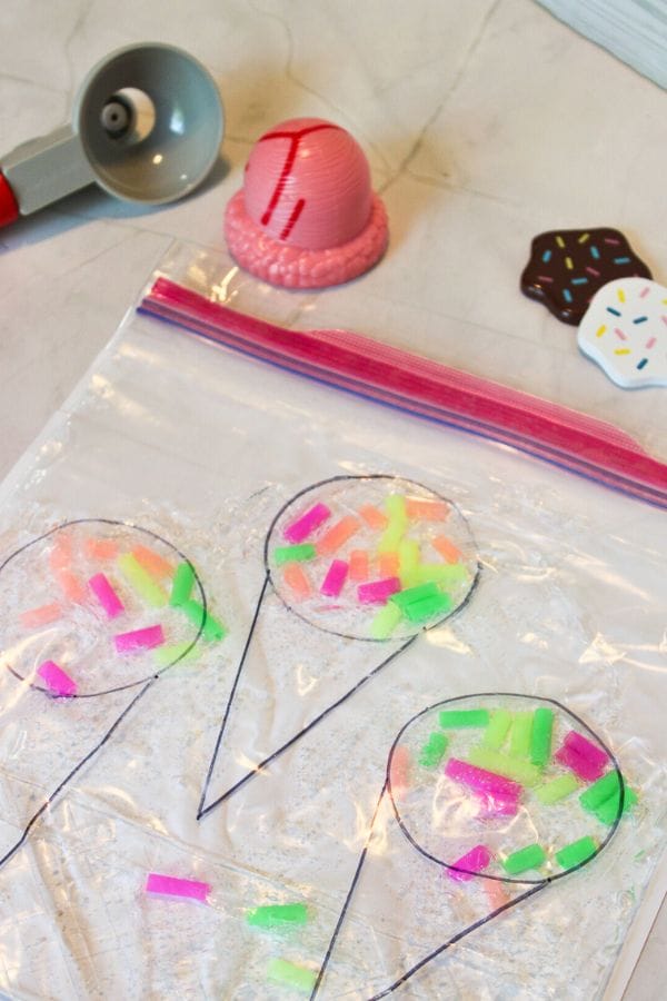 A Sprinkle Squish Bag adorned with ice cream cone drawings is filled with colorful sprinkles. Nearby, a scooper and pink scoop mold sit alongside ice cream-themed toys on the table, creating a whimsical dessert-inspired scene.