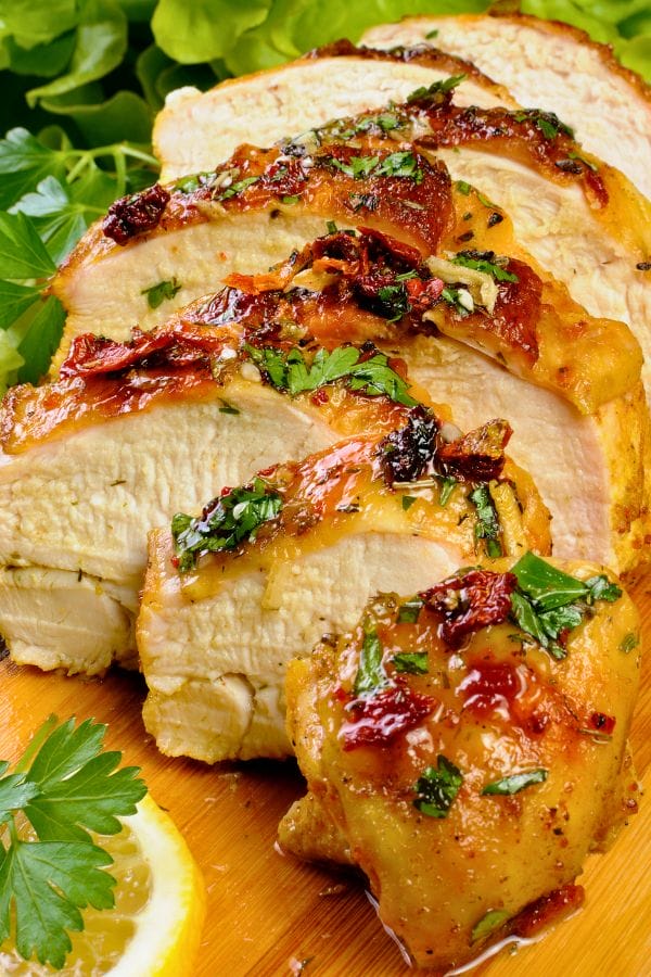 Sliced roasted chicken breast garnished with herbs and sun-dried tomatoes on a cutting board, with lemon and parsley in the background.