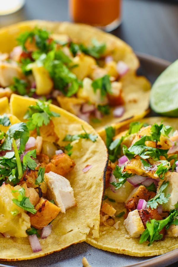 Close-up of three tacos filled with diced grilled chicken, pineapple, red onion, and cilantro on corn tortillas, garnished with lime wedges