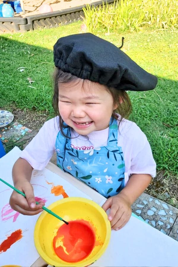 A child wearing a beret smiles as they celebrate their love for art, painting joyfully with a brush. They are outdoors, working with vibrant orange paint in a yellow bowl, set against the backdrop of a lush grassy area.