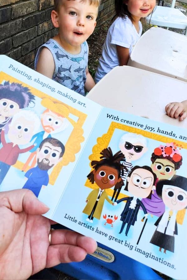 A person is holding an open children's book showcasing illustrations of diverse artists, capturing the essence of an art celebration for kids. In the background, two children sit at a white table, their eyes wide with curiosity and wonder.