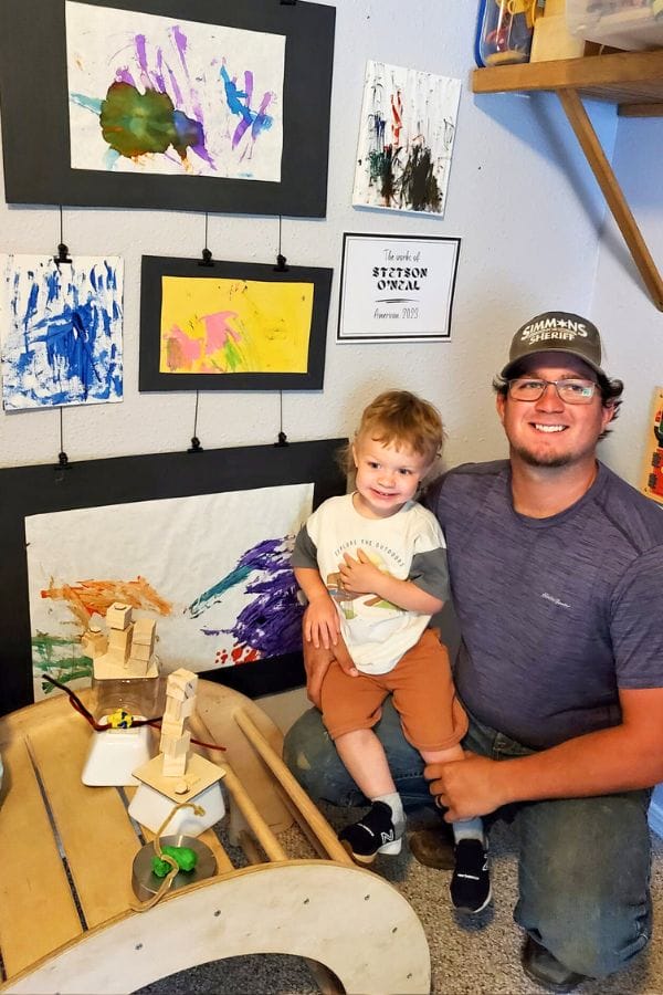 An adult and child pose by a wall displaying colorful children's artwork, capturing the spirit of an Art Celebration for Kids. The child smiles brightly, resting a hand on a small wooden sculpture.