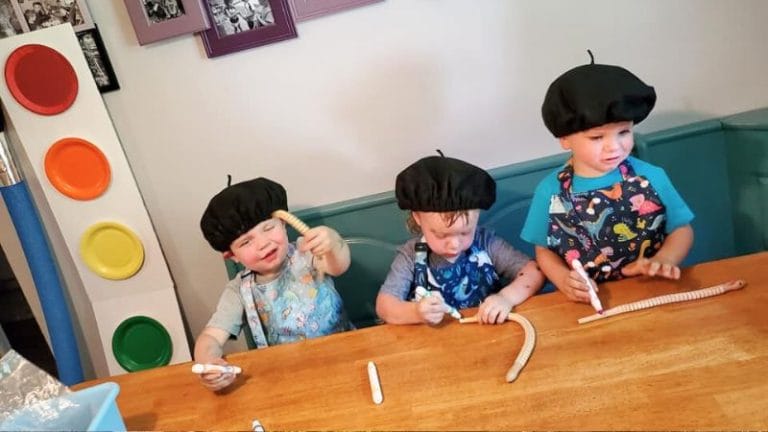 Three toddlers wearing black berets sit at a table, each holding white markers and playing with flexible tubes, in what seems like an art celebration for kids.
