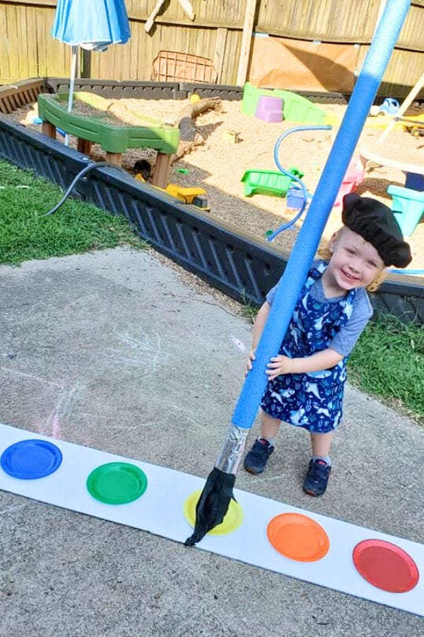 In a festive art celebration for kids, a child wearing a beret holds a large paintbrush prop near colorful paper circles, with a sandbox providing the perfect playful backdrop.
