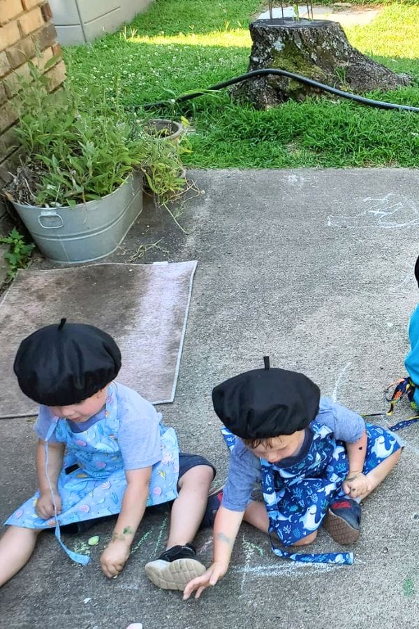 Two young children wearing berets sit on a concrete surface, joyfully celebrating art as they draw with chalk. Nearby, grass sways gently next to a metal tub filled with vibrant plants.