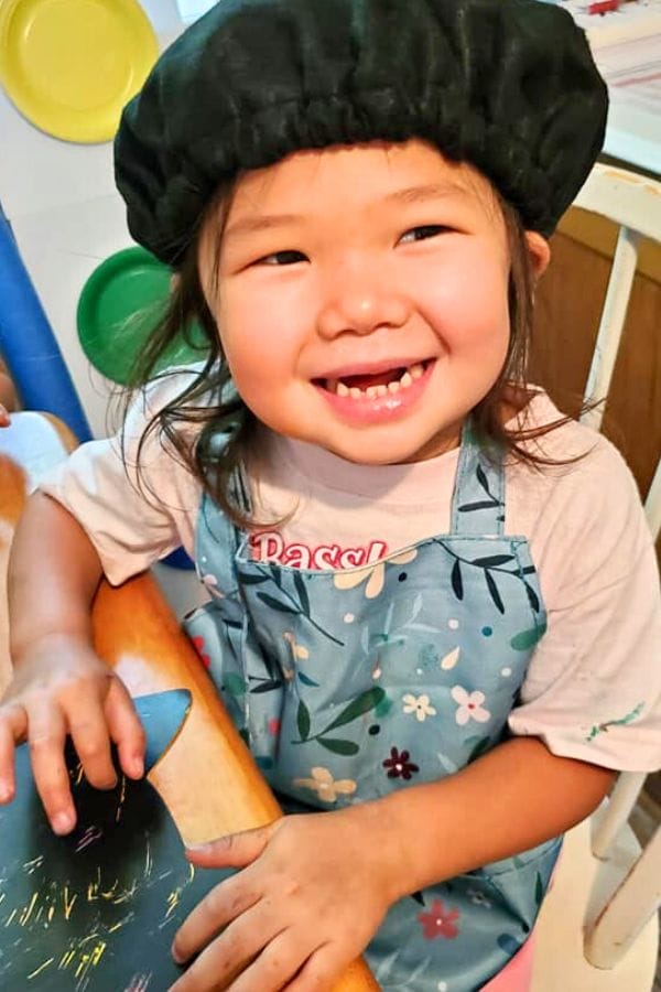 A smiling toddler, donned in a black chef hat and colorful apron, is the picture of joy at an art celebration for kids.