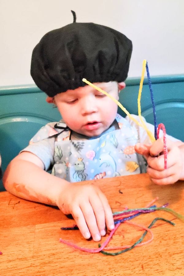 A toddler wearing a black beret is sitting at a table, holding colorful pipe cleaners and concentrating on them in what seems like an intimate art celebration for kids.
