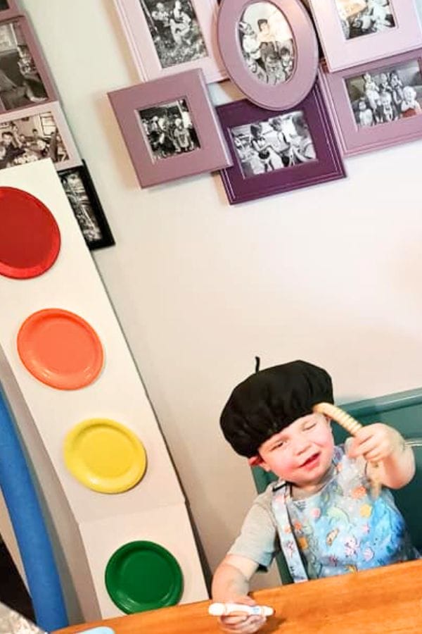 A child, donned in a black hat and apron, joyfully engages in an art celebration for kids, holding a paintbrush while surrounded by vibrant paint pots. The background showcases a collage of black and white photos in elegant frames.
