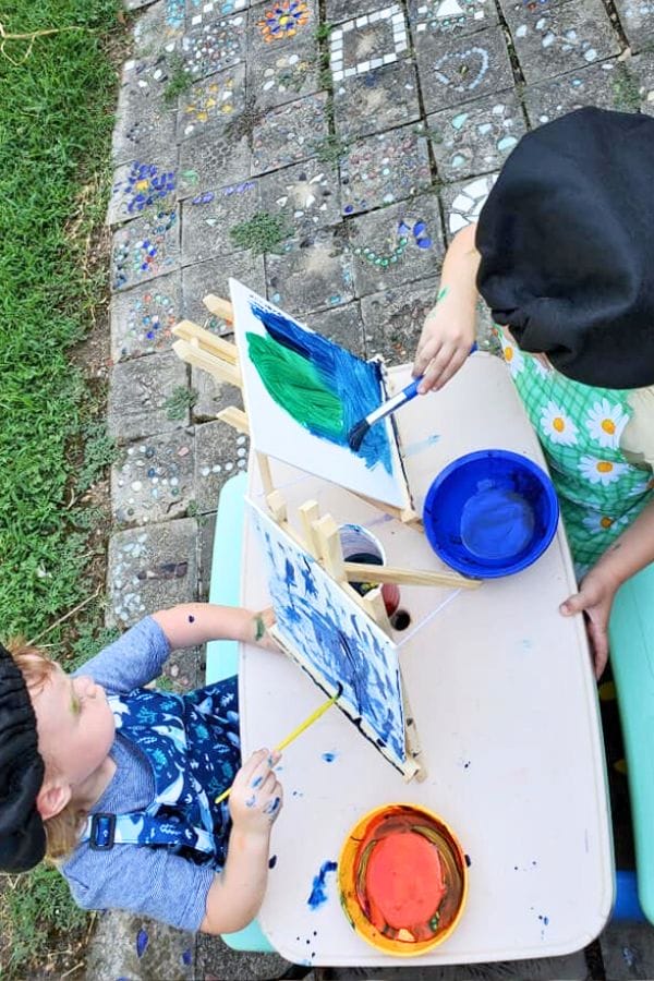 Two children happily painting on small easels outdoors, turning the day into an art celebration for kids. Dressed in aprons and splashing vibrant blue and green paints, they bring color to the ground adorned with charming tiles and stones.