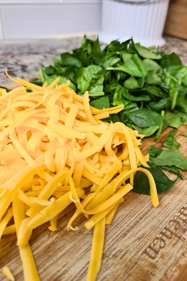 A wooden cutting board holds shredded cheddar cheese and chopped spinach, perfect for crafting a scrumptious Tuna Bacon and Mushroom Quiche.