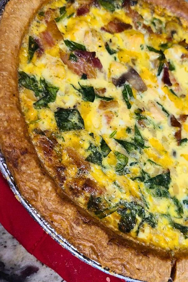 A close-up of a freshly baked Tuna Bacon and Mushroom Quiche with a golden brown crust, topped with visible pieces of spinach and cheese, resting on a vibrant red surface.