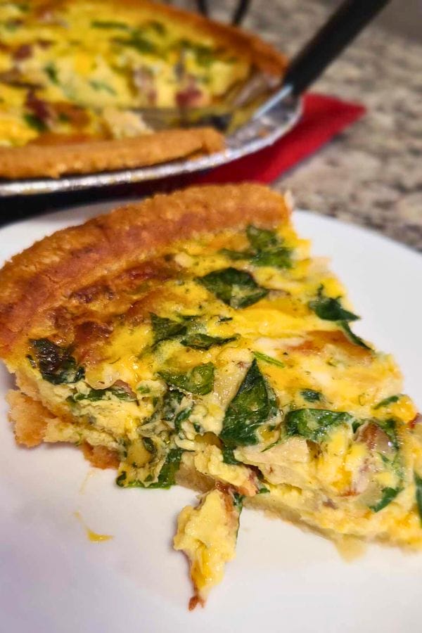 A slice of spinach quiche on a white plate pairs beautifully with the flavors from a Tuna Bacon and Mushroom Quiche resting in the background on a red cloth.
