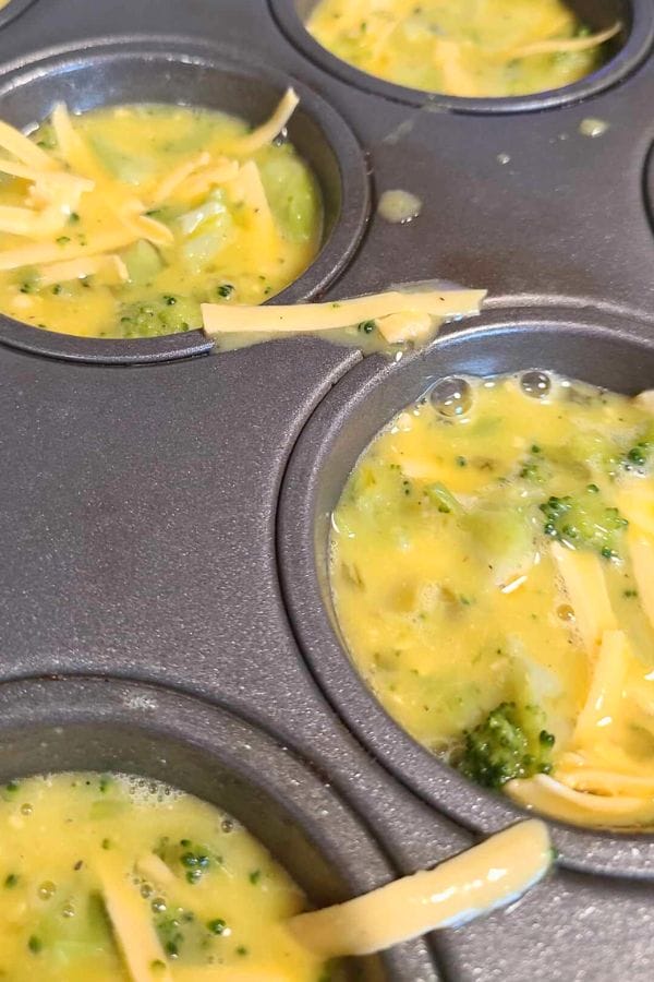 A muffin tray brimming with the delightful mix of Broccoli Cheese Egg Cups, featuring uncooked egg mixture, fresh broccoli, and a generous sprinkle of shredded cheese.