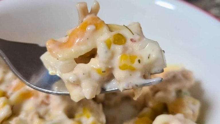 Close-up of a fork holding creamy Tuna and Corn Pasta Salad, with visible pieces of corn and other ingredients in a white bowl.