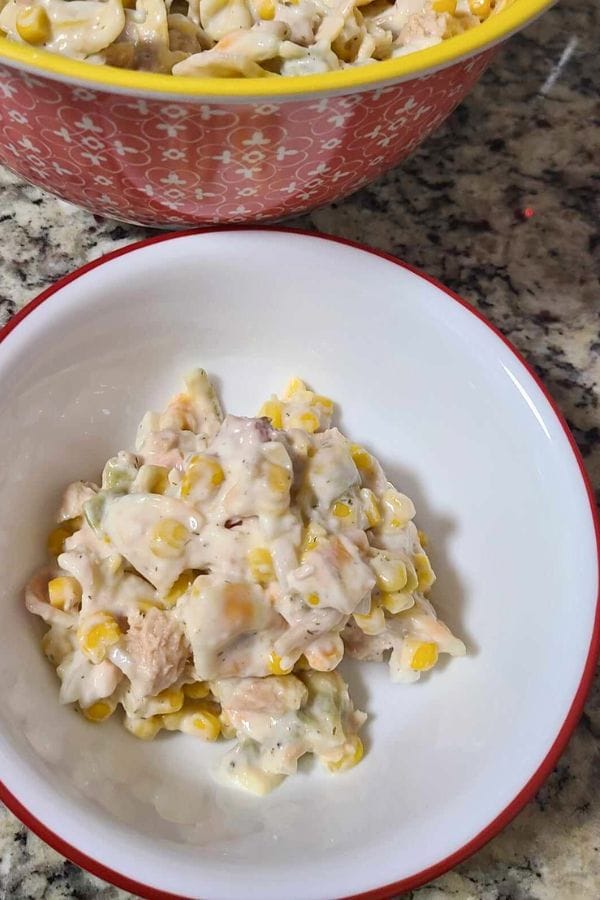 A plate of creamy tuna and corn pasta salad with shredded chicken is served on a red-rimmed white plate. In the background, a larger bowl with more salad is partially visible.