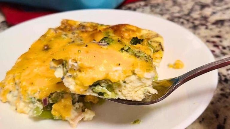 A fork lifts a bite of a cheesy broccoli and egg casserole from a white plate on a granite countertop, recalling the savory flavors of a crustless tuna quiche.