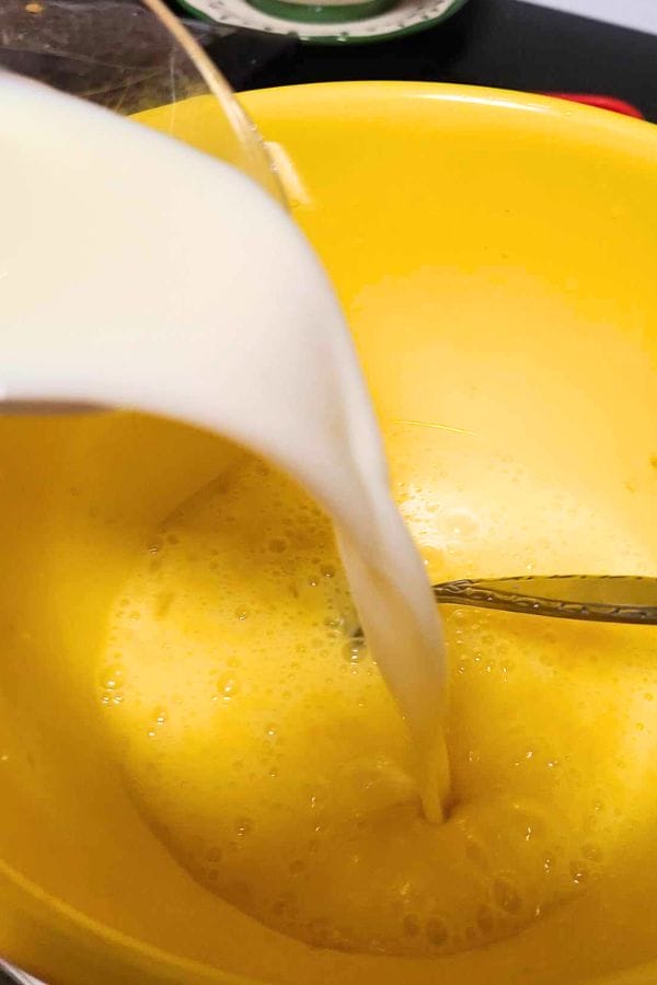 Milk being poured into a yellow bowl containing a whisked egg mixture, forming the base for a crustless tuna quiche.