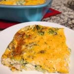 A slice of broccoli and cheese casserole graces a white plate, with the rest of the crustless tuna quiche-inspired dish in a blue baking pan in the background.