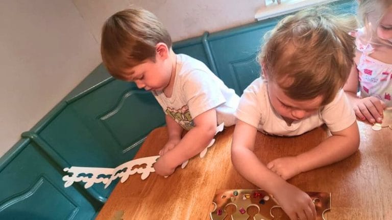 Two young children are engaged in crafting activities at a wooden table, using paper cutouts and small colorful beads, as part of a classic literature themed tea party.