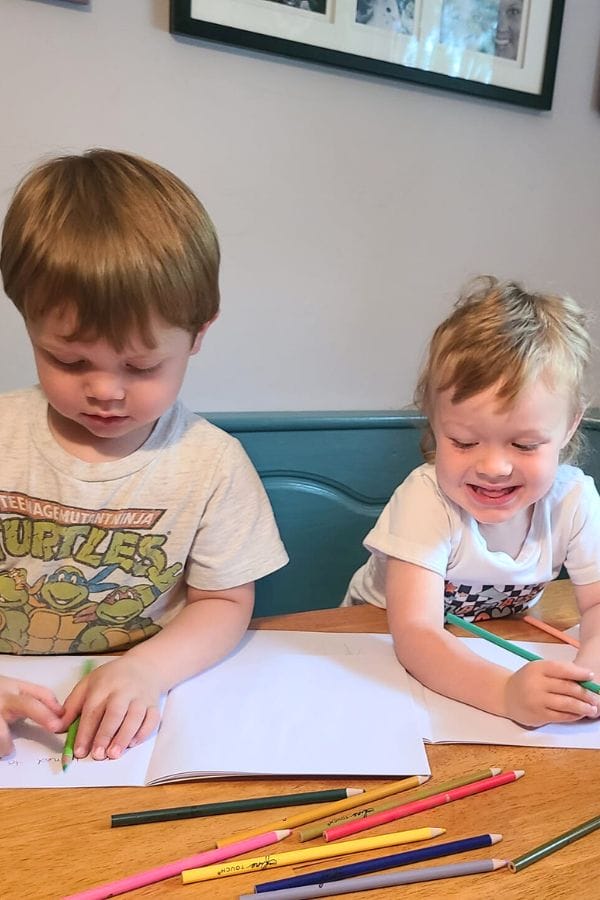 Two young children sit at a table, immersed in a classic literature themed tea party, as they draw with colored pencils on paper.