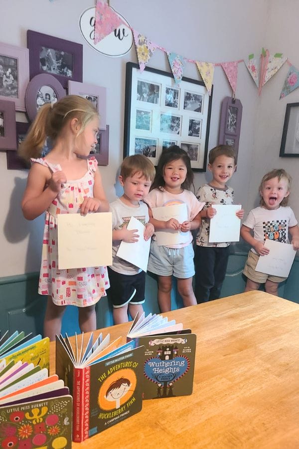 Five young children stand together, papers in hand and smiles on their faces, amidst a room adorned with framed photos and bunting. The table in the foreground displays books, setting the perfect scene for a classic literature-themed tea party.