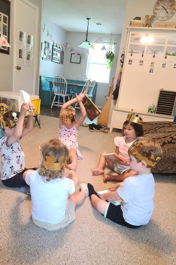 Five children wearing crown hats sit on the floor in a circle, each holding a book, as if at a classic literature themed tea party, with a dining area as their backdrop.