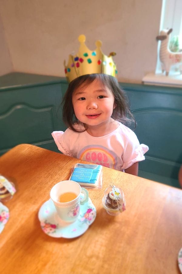 A young child wearing a crown beams at a table set for a classic literature-themed tea party, where a cup of tea, a cupcake, and a blue book await.