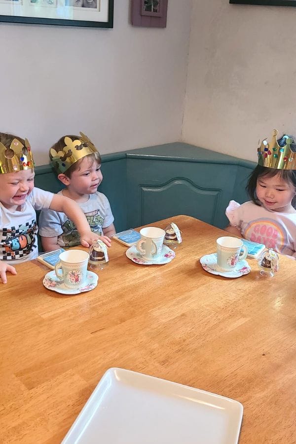 Three children wearing paper crowns sit at a table with teacups and snacks, engaging in a Classic Literature Themed Tea Party. They smile and interact, bringing stories to life with their imaginations.